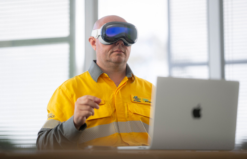 Un employé porte l’Apple Vision Pro et effectue un mouvement de pince avec ses doigts, tandis qu’un MacBook Pro argent est ouvert sur le bureau